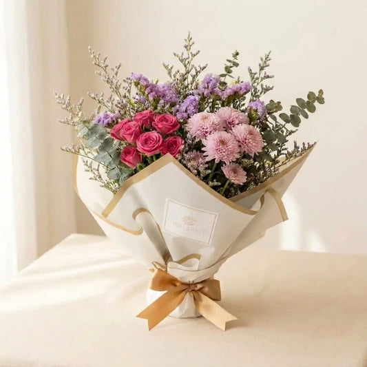 Mini ramo Florencia con rosas fucsia, pompones rosados y statice lila envuelto en papel blanco con moño dorado