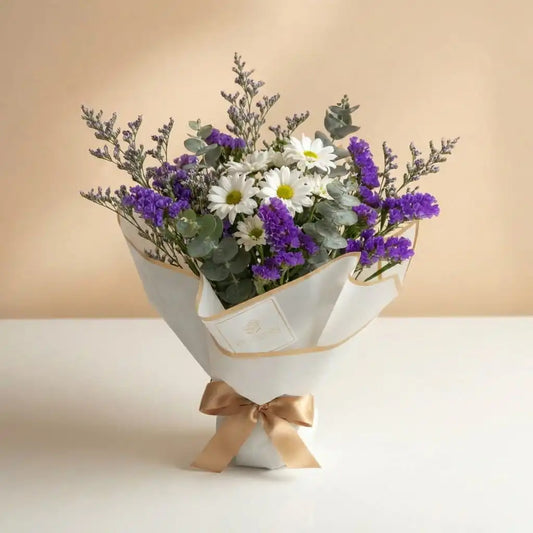 Ramo de flores elegantes con margaritas blancas, estatice morado y eucalipto envuelto en papel premium con lazo dorado de Fruit Santé.